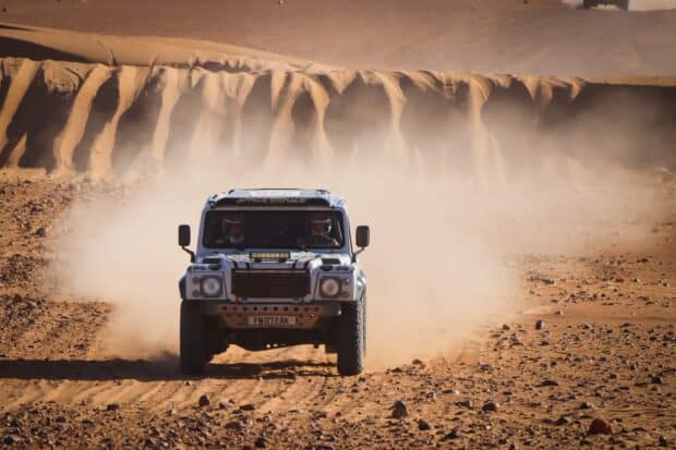 A rally raid vehicle is driving through a dusty desert trail with rocky terrain, HD Desktop Wallpaper