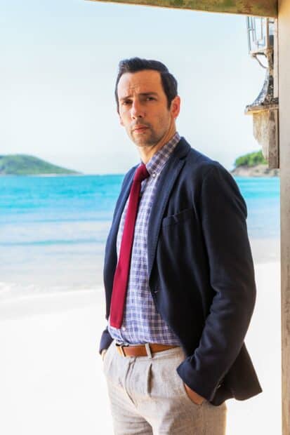 A detective from Death In Paradise TV Series stands confidently near the ocean shore, 2K Phone Wallpaper