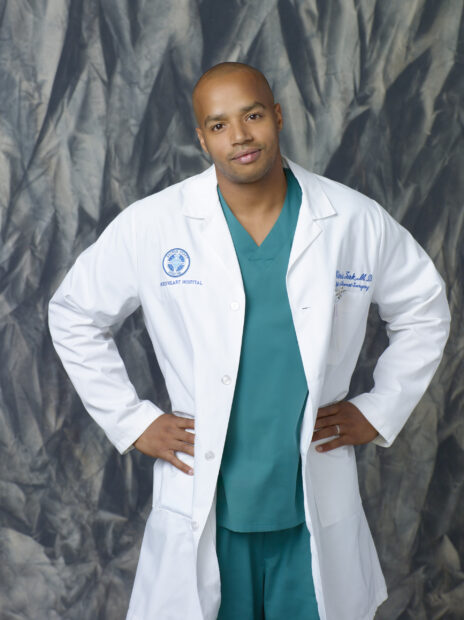 Donald Faison wearing a white coat and teal scrubs posing confidently indoors, 2K Desktop Wallpaper