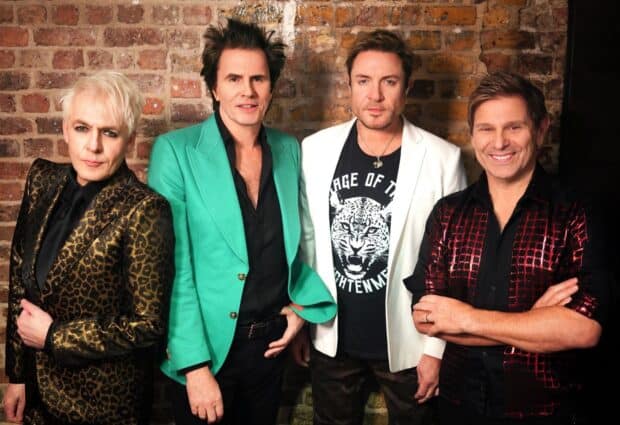 The four members of Duran Duran stand against a brick wall posing confidently in stylish outfits, HD Desktop Wallpaper