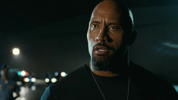 Dwayne Johnson looks focused and serious in a dimly lit scene with blurred lights behind him, HD Desktop Wallpaper