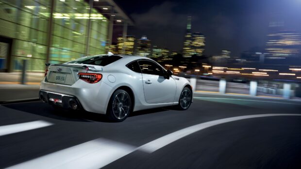 A white Subaru Brz drives rapidly on an urban road at night with city lights in the background, 4K Desktop Wallpaper