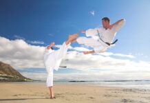 A karate practitioner performs a flying kick over another on a sandy shore under a blue sky, HD Desktop Wallpaper