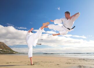 A karate practitioner performs a flying kick over another on a sandy shore under a blue sky, HD Desktop Wallpaper