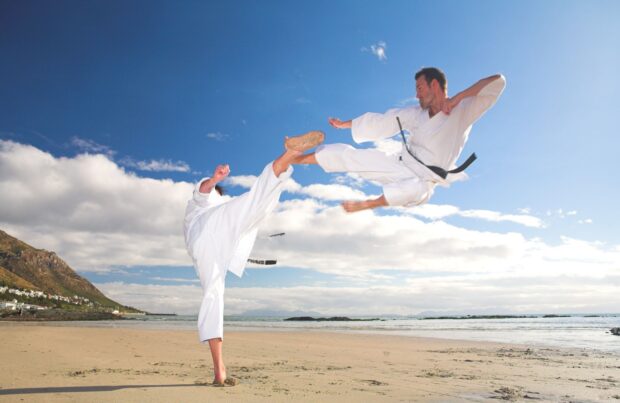 A karate practitioner performs a flying kick over another on a sandy shore under a blue sky, HD Desktop Wallpaper