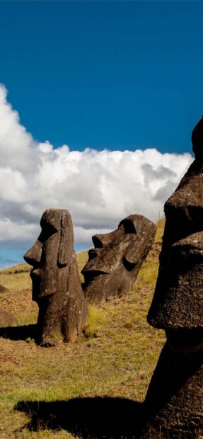 The Easter Island stone statues stand tall on a grassy hill under a blue sky with white clouds, 2K Phone Wallpaper