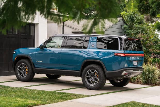 A blue Rivian electric SUV is parked outside a house surrounded by greenery, 2K Desktop Wallpaper