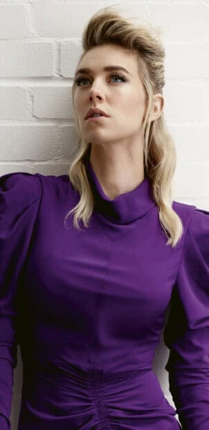Vanessa Kirby looks thoughtfully away in a purple dress against a white brick wall, 2K Mobile Wallpaper