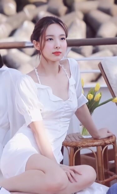 A graceful woman wearing a white dress sits beside a wooden table with flowers in soft natural light featuring Nayeon Wallpapers, HD Phone Wallpaper