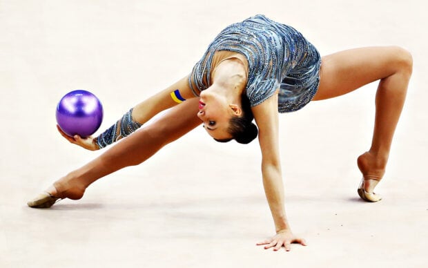 A rhythmic gymnast performs a flexible move holding a purple ball during a competition, HD Desktop Wallpaper
