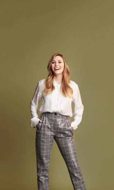 Elizabeth Olsen smiles confidently wearing a white blouse and plaid pants against a plain background