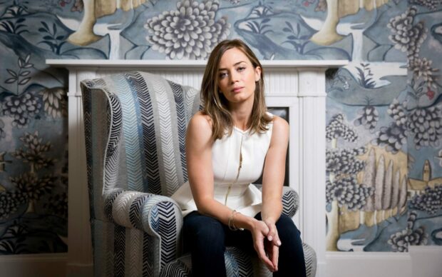 Emily Blunt is sitting thoughtfully on a patterned armchair with floral wallpaper in the background, HD Desktop Wallpaper