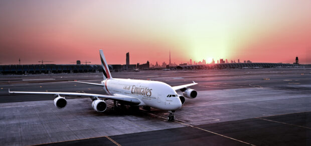 An Emirates Airbus A380 is parked on the runway at sunset with a city skyline in the background, 2K Desktop Wallpaper