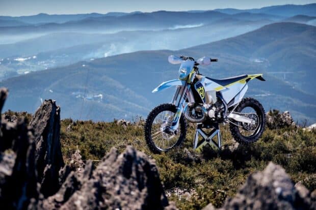 A white and blue enduro motorbike stands on rocky mountain terrain with distant hills in the background