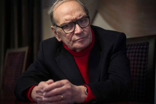 An elderly man wearing glasses and a red sweater is sitting thoughtfully in a dim room, Ennio Morricone Wallpapers