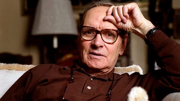 Ennio Morricone is sitting on a couch with a hand on his forehead in a relaxed setting, HD Desktop Wallpaper