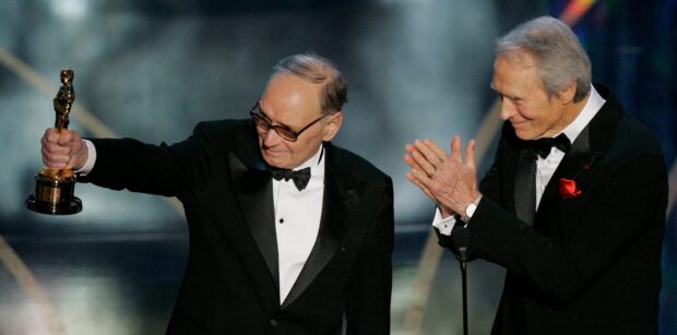 Ennio Morricone is holding a trophy while a man in a tuxedo applauds him on stage, HD Desktop Wallpaper