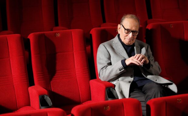 Ennio Morricone is sitting alone in a red theater with a thoughtful expression, 2K Desktop Wallpaper