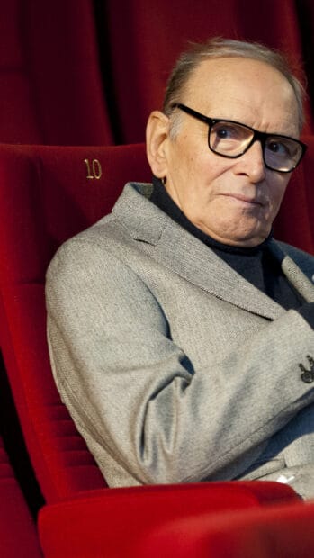 Ennio Morricone is wearing glasses and a grey jacket while sitting in a red theater seat, 2K Phone Wallpaper