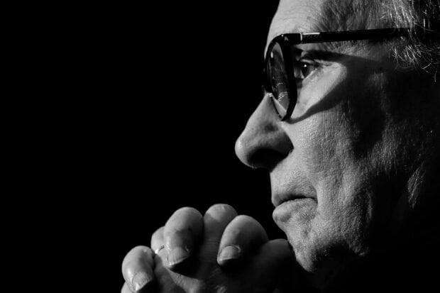 A close up black and white image of Ennio Morricone deep in thought with hands clasped, 2K Desktop Wallpaper