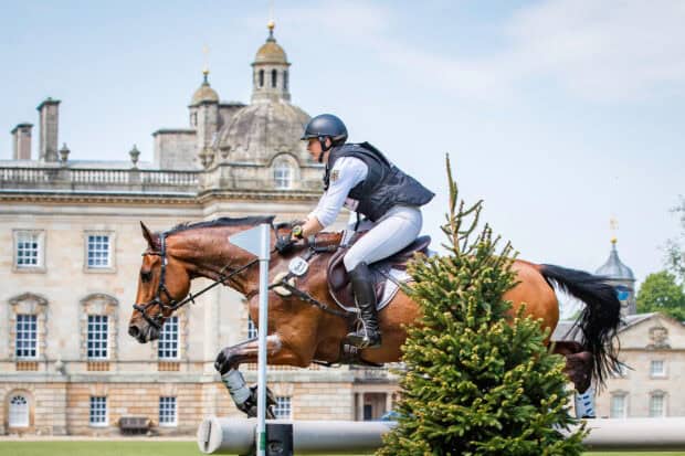 A rider in eventing gear jumps a horse over an obstacle near a historic building, HD Desktop Wallpaper