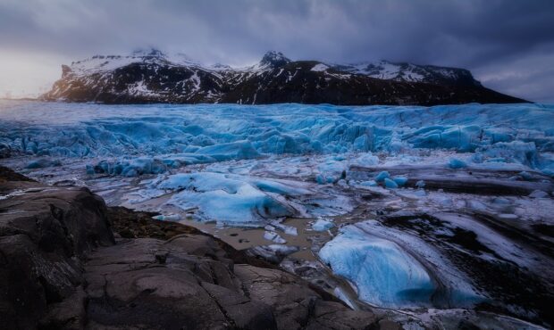 A vast glacier with blue ice stretches before dark rocky mountains under a cloudy sky, 2K Desktop Wallpaper