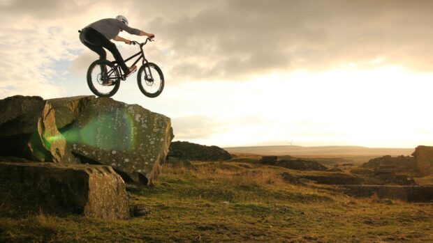 A BMX rider performs a stunt over large rocks in a rugged landscape, HD Desktop Wallpaper