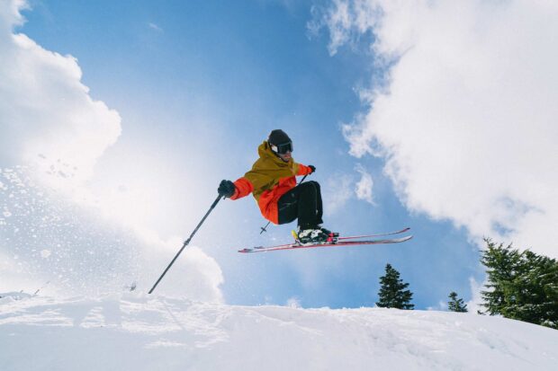 A skier wearing orange and yellow performs a high jump against a blue sky with clouds, HD Desktop Wallpaper