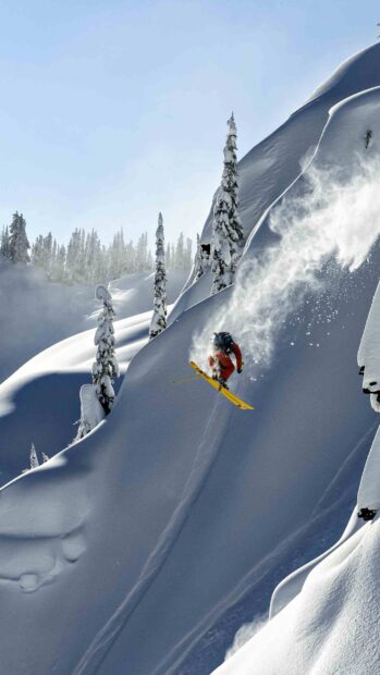 A skier performs a jump on a steep snowy mountain surrounded by snow covered trees in fresh powder, 2K Phone Wallpaper