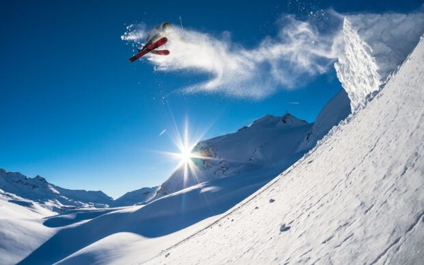 A skier is performing a jump creating a spray of snow on a sunny mountain slope, HD Desktop Wallpaper