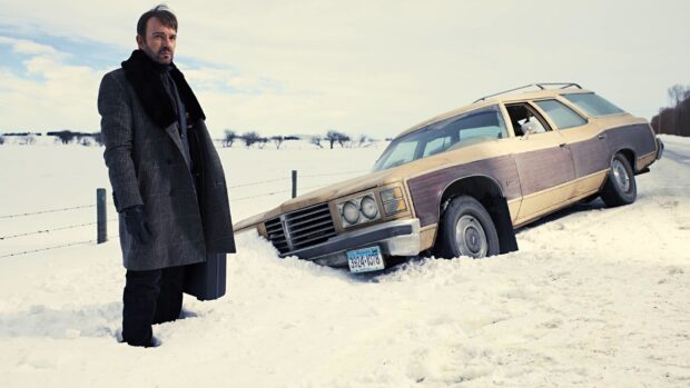 A man stands in deep snow next to a car stuck in the snow in a Fargo scene, 2K Desktop Wallpaper