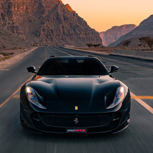 A black Ferrari 812 Gts is driving on a mountain road during sunset with rocky cliffs in the background