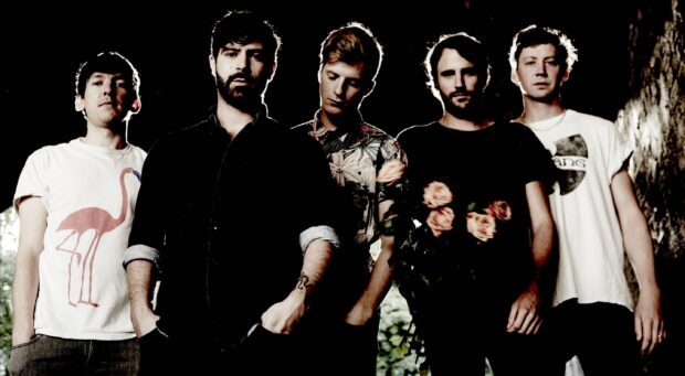 The Foals band members stand together in a dark environment with soft natural light behind them, 2K Desktop Wallpaper