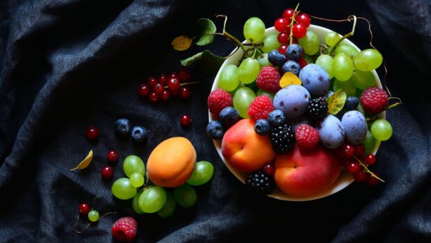 A bowl filled with apricots, grapes, berries, and plums on dark fabric featuring apricot wallpapers, 4K Desktop Wallpaper