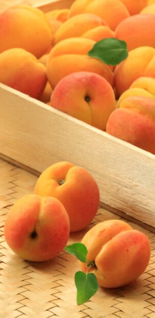 A group of apricots with green leaves resting on a woven surface near a wooden crate, 2K Mobile Wallpaper
