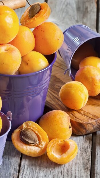 Fresh apricots in purple buckets and on a rustic wooden surface with one cut open showing the seed, 2K Mobile Wallpaper