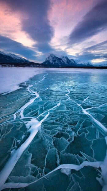 A frozen lake with cracked ice leads toward snow covered mountains in glacier wallpapers, 4K Phone Wallpaper