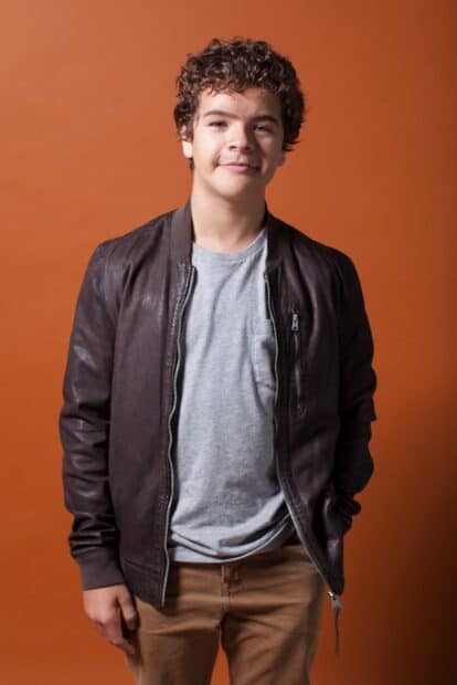 Gaten Matarazzo stands confidently against an orange background wearing a brown jacket, HD Mobile Wallpaper