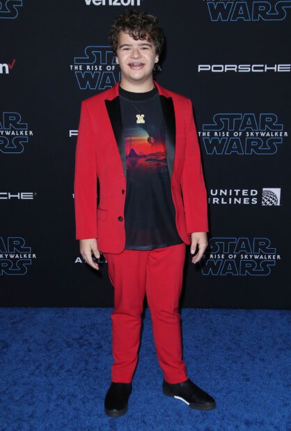 Gaten Matarazzo wearing a red suit with a graphic t shirt standing on a blue carpet at a Star Wars event, HD Desktop Wallpaper