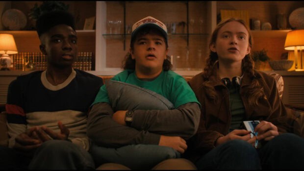 Gaten Matarazzo sits on a couch between two friends in a warmly lit room, HD Desktop Wallpaper
