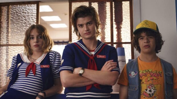 Gaten Matarazzo stands with crossed arms between two friends indoors, 4K Desktop Wallpaper