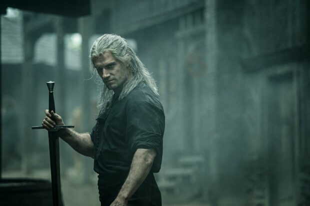 Geralt stands solemnly gripping his sword in a misty, ancient village scene from The Witcher Tv Series, HD Desktop Wallpaper