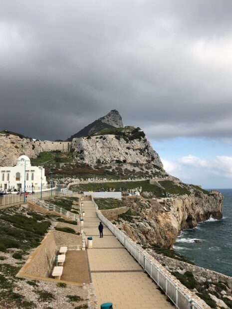 A person walking along the paved path near the rocky cliffs in Gibraltar with cloudy skies, HD Phone Wallpaper
