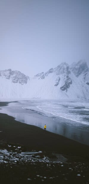 A person in a yellow jacket walks along the dark beach near glacier snowy mountains, HD Phone Wallpaper