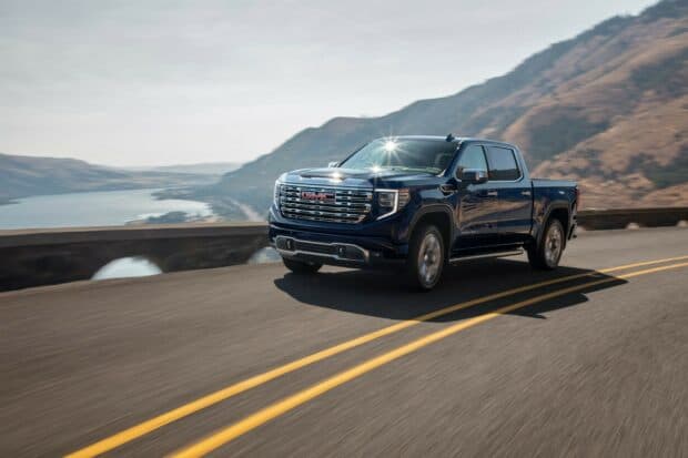 A blue Gmc Sierra is driving along a curved mountain road with water and hills in the background, HD Desktop Wallpaper