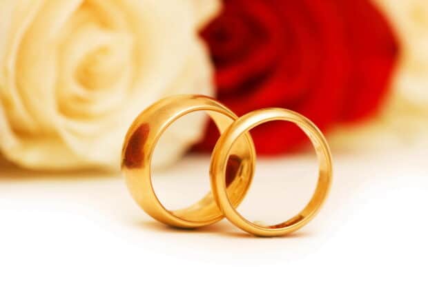 Two golden wedding rings placed on a white surface with red and white roses behind them, 2K Desktop Wallpaper