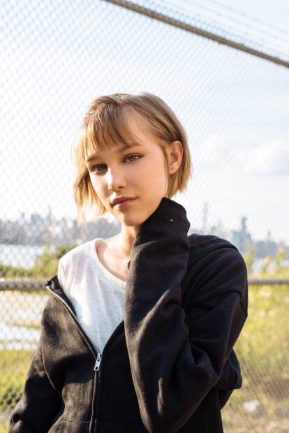 Grace Vanderwaal looks thoughtfully at the camera wearing a black jacket near a fence, HD Mobile Wallpaper