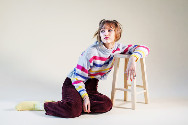 A young woman wearing colorful striped sweater sits on the floor beside a wooden stool looking thoughtfully, HD Desktop Wallpaper