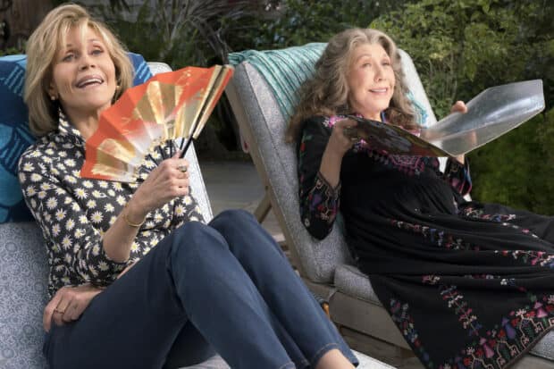 Grace and Frankie relaxing on lounge chairs with a fan and a book in a garden, HD Desktop Wallpaper