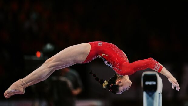 A gymnast in a red outfit performs a balanced flip on the beam during a competition, HD Desktop Wallpaper